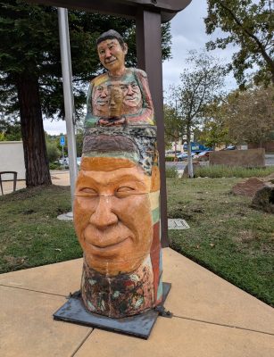 A stacked ceramic sculture about six foot tall. The bottom portion is a large, brown relief of a face, with the upper part being a figure's torso. The whole piece is decorated in many colors.