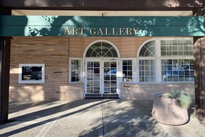 Picture of the storefront of the gallery with a wide green sign and golden text "ART GALLERY" on it. the doors are with covered with square windows.