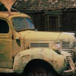 Gallery 3 - image of a rusty old yellow car on the left and an old wooden cabin on right side in the background