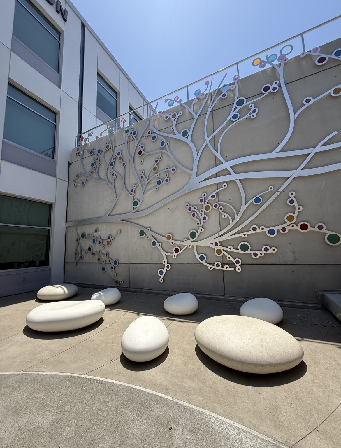 Gallery 3 - A 19-foot-high outdoor sculpture made of aluminum and illuminated with soft LED lights, featuring upward-reaching, branch-like forms inspired by the human nervous system and local plant life. A few large stone shaped benches in front of it.