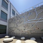 Gallery 3 - A 19-foot-high outdoor sculpture made of aluminum and illuminated with soft LED lights, featuring upward-reaching, branch-like forms inspired by the human nervous system and local plant life. A few large stone shaped benches in front of it.