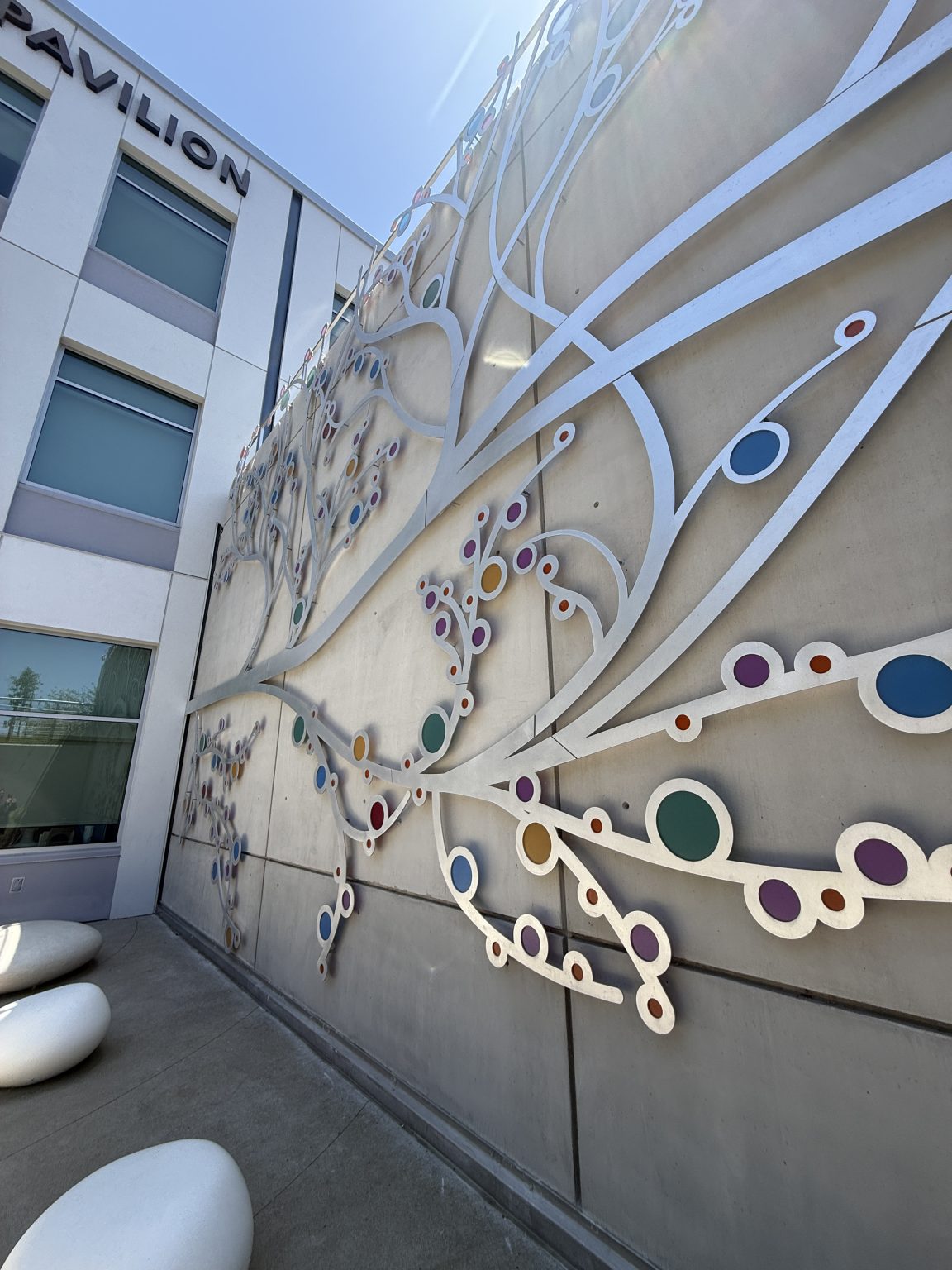Gallery 5 - A 19-foot-high outdoor sculpture made of aluminum and illuminated with soft LED lights, featuring upward-reaching, branch-like forms inspired by the human nervous system and local plant life. A few large stone shaped benches in front of it.