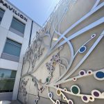 Gallery 5 - A 19-foot-high outdoor sculpture made of aluminum and illuminated with soft LED lights, featuring upward-reaching, branch-like forms inspired by the human nervous system and local plant life. A few large stone shaped benches in front of it.