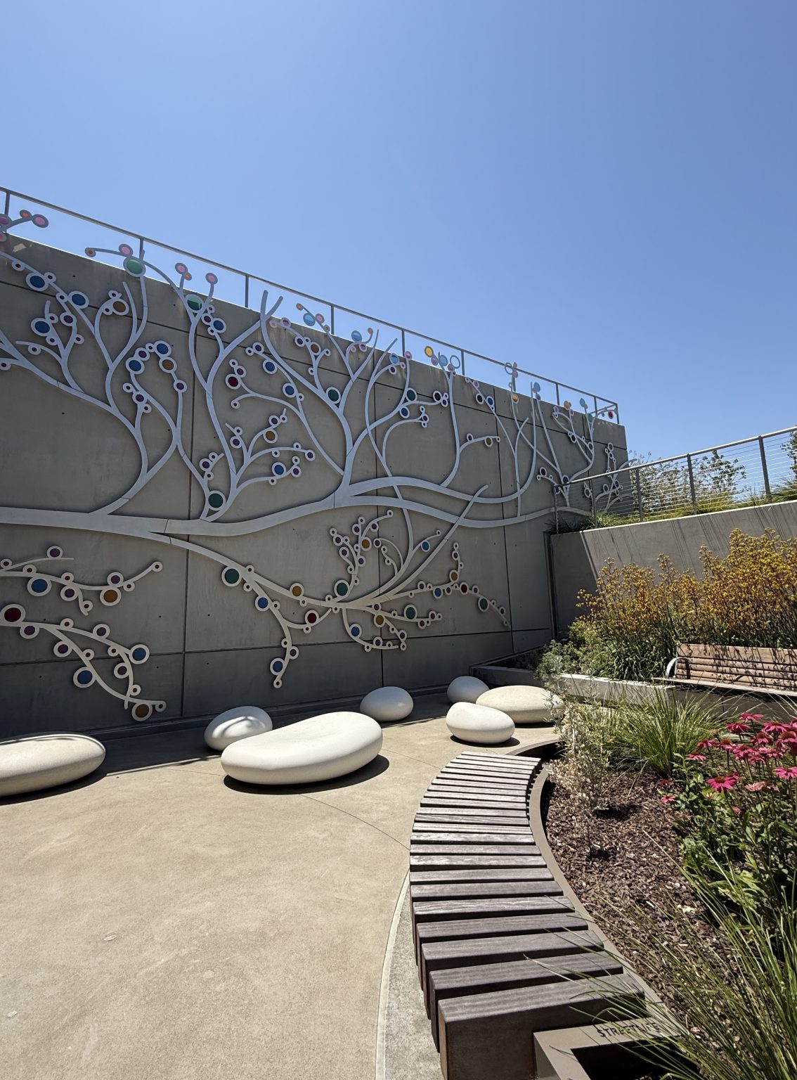 Gallery 8 - A 19-foot-high outdoor sculpture made of aluminum and illuminated with soft LED lights, featuring upward-reaching, branch-like forms inspired by the human nervous system and local plant life. A few large stone shaped benches in front of it.