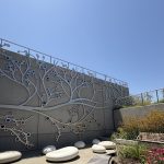 Gallery 8 - A 19-foot-high outdoor sculpture made of aluminum and illuminated with soft LED lights, featuring upward-reaching, branch-like forms inspired by the human nervous system and local plant life. A few large stone shaped benches in front of it.