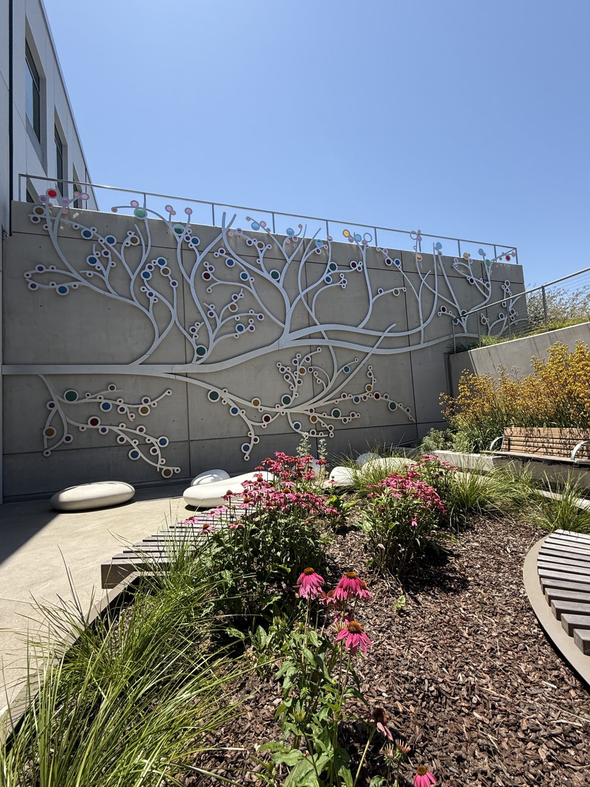 Gallery 9 - A 19-foot-high outdoor sculpture made of aluminum and illuminated with soft LED lights, featuring upward-reaching, branch-like forms inspired by the human nervous system and local plant life. A few large stone shaped benches in front of it.