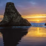 Gallery 1 - Two large sea stacks stand in the ocean under a sunset sky. The sun, a bright point of light, is low on the horizon between the two rock formations. The sky displays colors of orange, yellow, and purple. The water in the foreground reflects these sky colors, and small waves are visible around the base of the sea stacks.