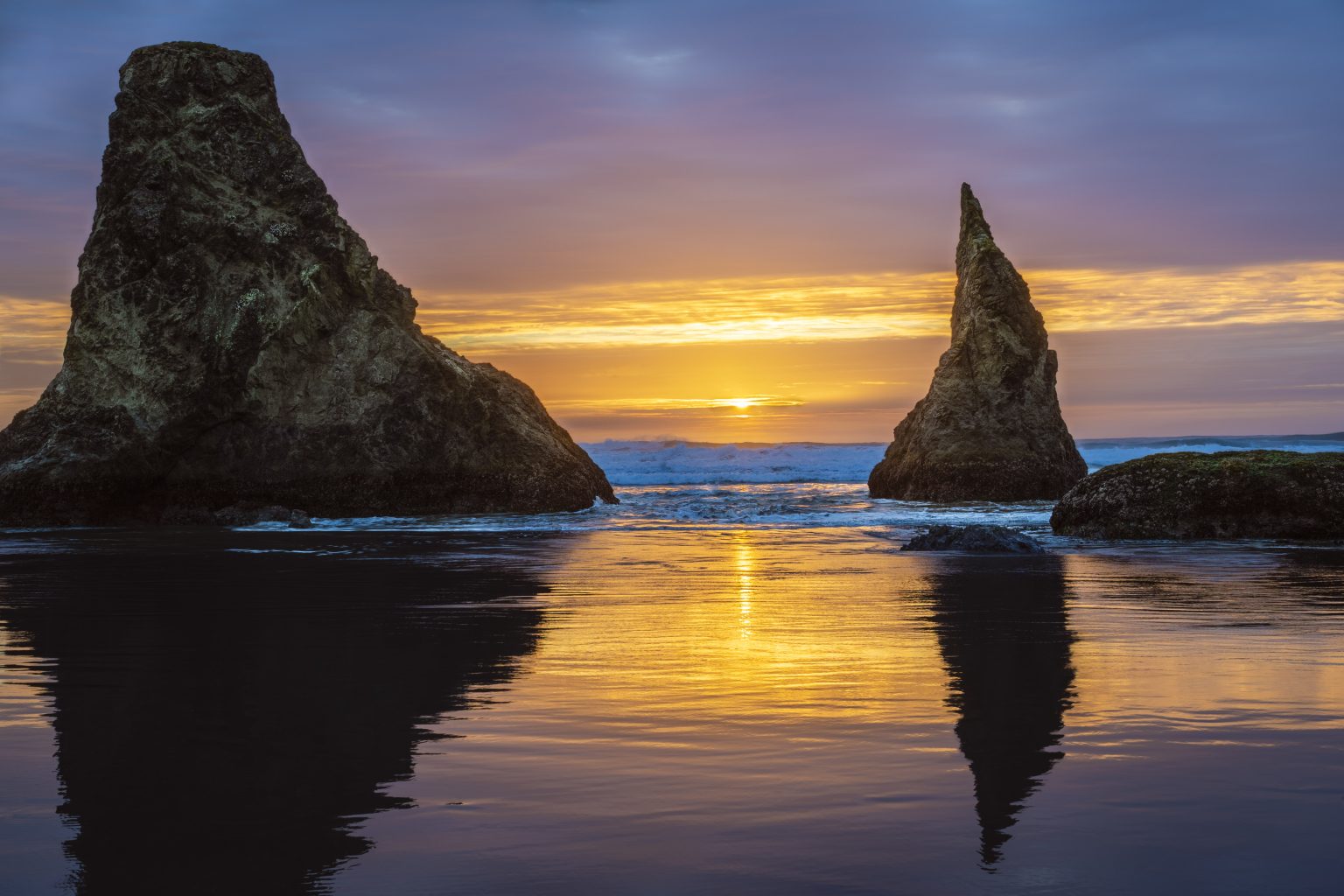 Gallery 1 - Two large sea stacks stand in the ocean under a sunset sky. The sun, a bright point of light, is low on the horizon between the two rock formations. The sky displays colors of orange, yellow, and purple. The water in the foreground reflects these sky colors, and small waves are visible around the base of the sea stacks.