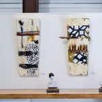 Gallery 2 - Three tall, narrow mixed-media artworks hang on a white wall above a wooden surface. The artworks have a textured, off-white background with black, abstract patterns. They are adorned with various materials like yarn, small beads, and what looks like hair or fur. Two small, white bust sculptures sit on the wooden surface beneath the artworks.
