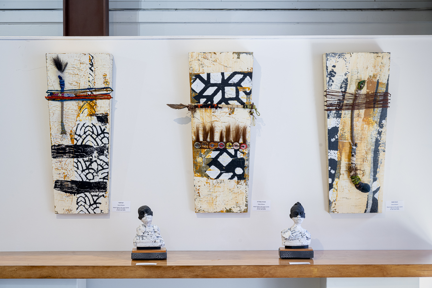 Gallery 2 - Three tall, narrow mixed-media artworks hang on a white wall above a wooden surface. The artworks have a textured, off-white background with black, abstract patterns. They are adorned with various materials like yarn, small beads, and what looks like hair or fur. Two small, white bust sculptures sit on the wooden surface beneath the artworks.