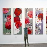 Gallery 3 - A long, white gallery wall is adorned with six tall, narrow paintings featuring abstract red floral shapes on backgrounds of gray, white, and purplish tones. The paintings are hung in a line, creating a single, continuous display. In the foreground, a tall, slender sculpture of a human torso stands on the floor, and to the right, a wooden counter holds several small busts.