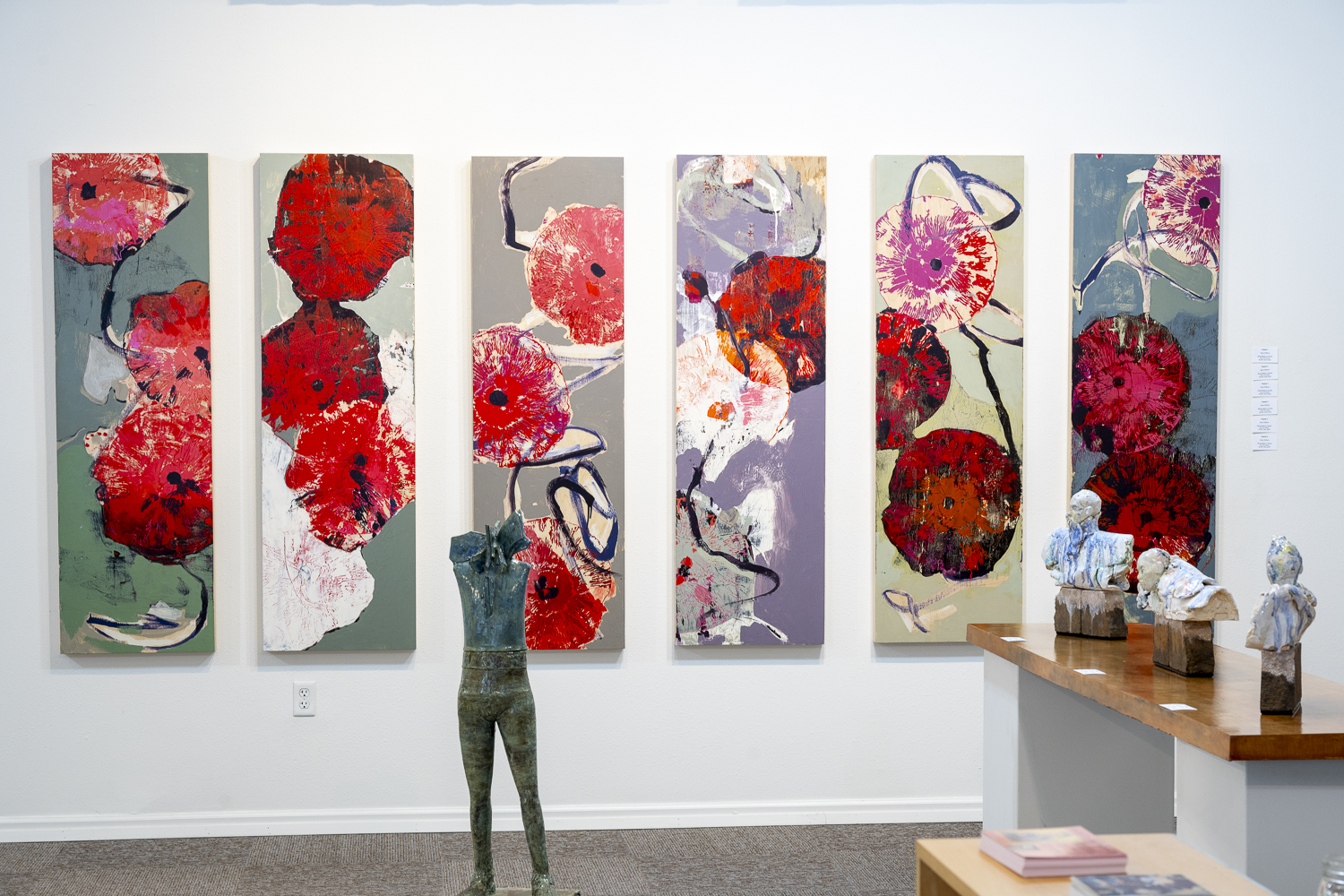 Gallery 3 - A long, white gallery wall is adorned with six tall, narrow paintings featuring abstract red floral shapes on backgrounds of gray, white, and purplish tones. The paintings are hung in a line, creating a single, continuous display. In the foreground, a tall, slender sculpture of a human torso stands on the floor, and to the right, a wooden counter holds several small busts.