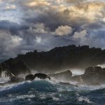 Gallery 3 - A dark, rocky outcrop in the ocean is being hit by large waves, creating a mist of sea spray. The rocks are dark and jagged, with some lighter patches visible. The sky above is filled with dramatic, dark clouds with patches of sunlight breaking through. Two birds are silhouetted on the ridge of the rocks.