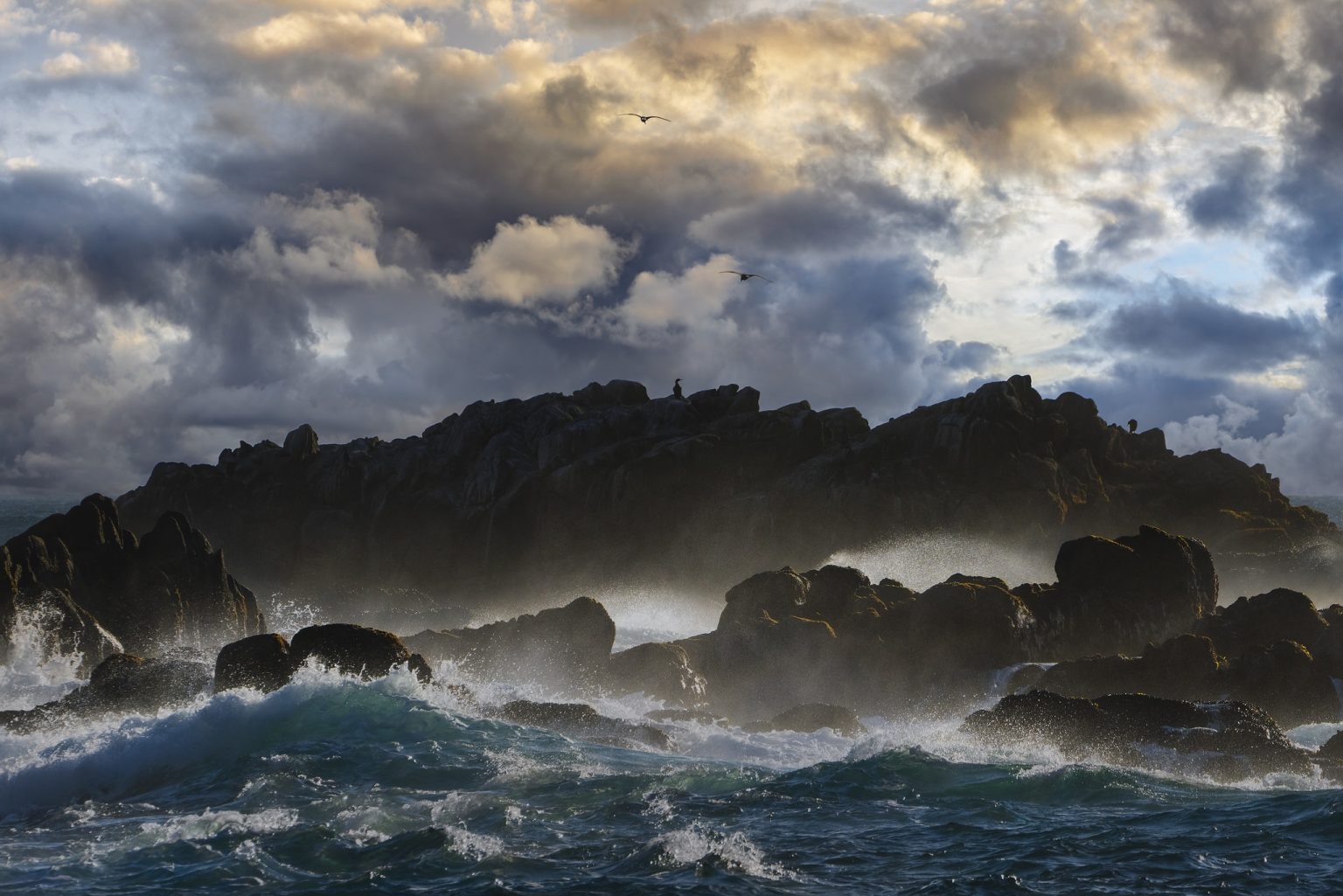 Gallery 3 - A dark, rocky outcrop in the ocean is being hit by large waves, creating a mist of sea spray. The rocks are dark and jagged, with some lighter patches visible. The sky above is filled with dramatic, dark clouds with patches of sunlight breaking through. Two birds are silhouetted on the ridge of the rocks.