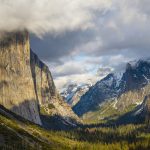 Gallery 4 - A scenic view of Yosemite Valley, with prominent rock formations on either side. On the left is the sheer face of El Capitan, partly illuminated by sunlight and partly shrouded in clouds. On the right, Bridalveil Fall cascades down a cliff face. The valley floor is covered in a dense forest of evergreen trees, with snowy mountains visible in the distance. The sky is partially cloudy, with some sunlight breaking through.
