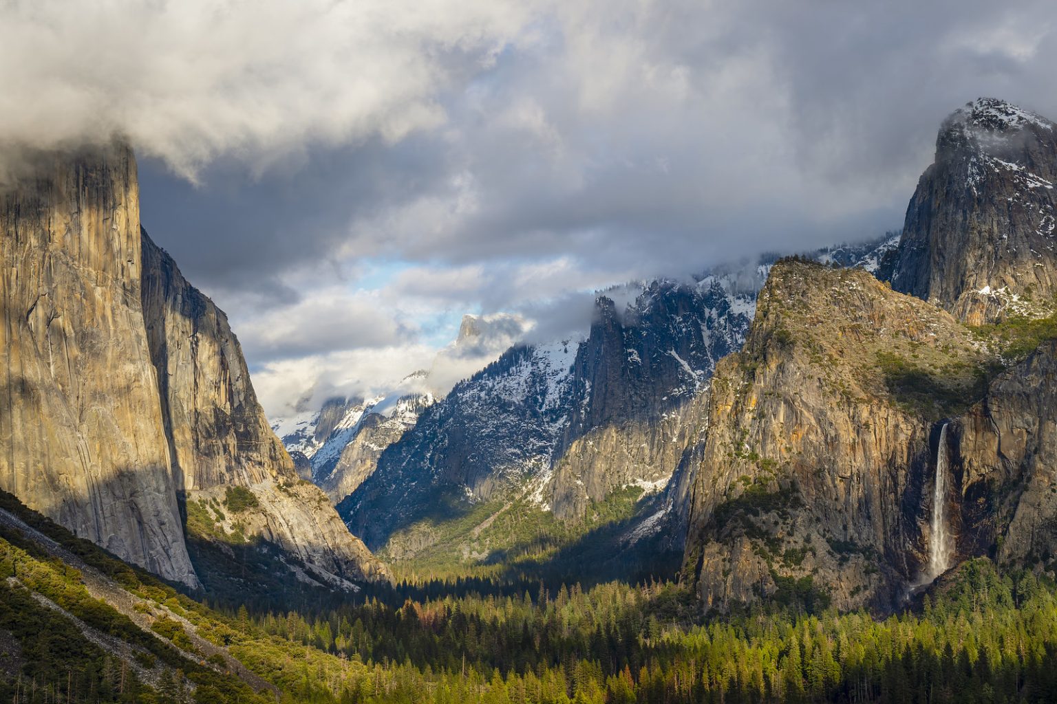Gallery 4 - A scenic view of Yosemite Valley, with prominent rock formations on either side. On the left is the sheer face of El Capitan, partly illuminated by sunlight and partly shrouded in clouds. On the right, Bridalveil Fall cascades down a cliff face. The valley floor is covered in a dense forest of evergreen trees, with snowy mountains visible in the distance. The sky is partially cloudy, with some sunlight breaking through.