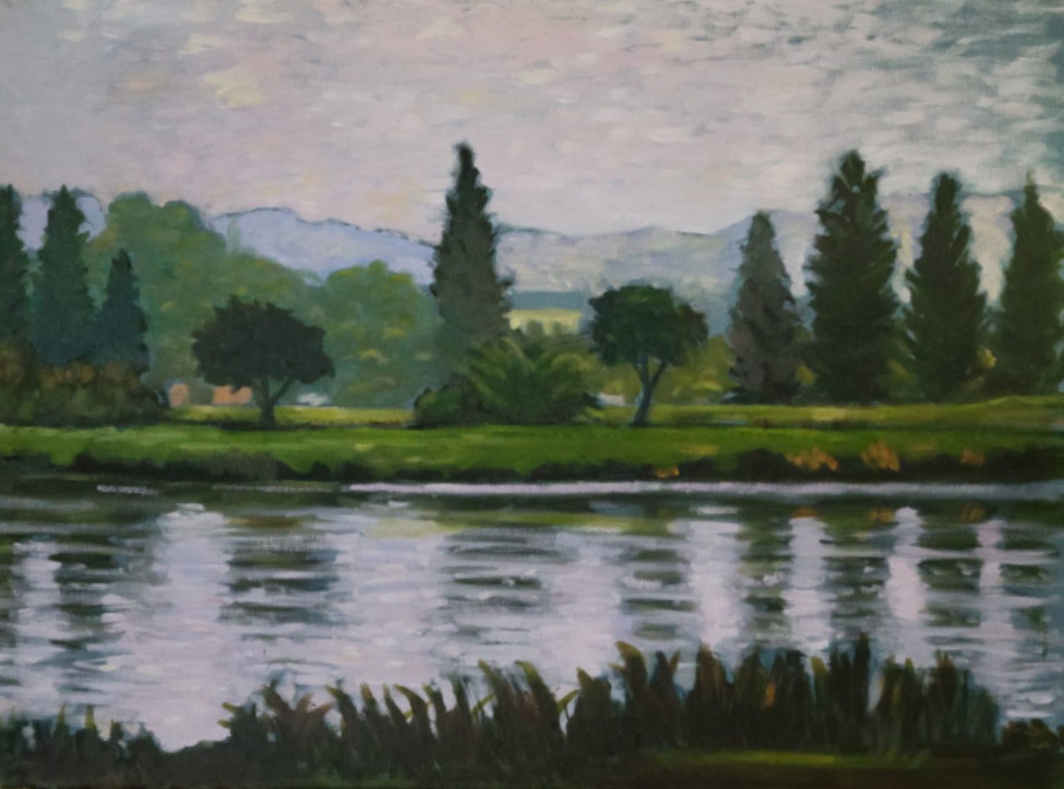 Gallery 4 - Water and reflections. A landscape painting of a lake or river reflecting the shore. The foreground is a body of water with rippling, dark blue and grey reflections of the sky and trees. The closest part of the bank has thick, dark green grass and reeds. The middle ground features a variety of lush green trees, including some tall, thin evergreens and rounder deciduous trees. A grassy green hill and a building are visible behind the trees, with a range of soft, hazy blue-grey mountains in the distance. The sky is a pale, cloudy grey. The artist's visible brushstrokes give the painting a textured, impressionistic feel.