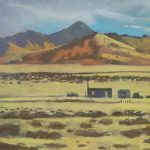 Gallery 5 - Small house in the desert. A painting of a vast, open desert landscape. The foreground is a light tan and gold field with scattered dark patches of shrubs. In the middle ground, three small, dark purple or grey structures sit alone, surrounded by a wire fence. One building has a distinctly steep roof, and another has a chimney. The structures are dwarfed by a distant range of large, rolling mountains with warm orange, yellow, and brown tones. A cloudy blue sky is above the mountains. The painting has a soft, chalky texture.