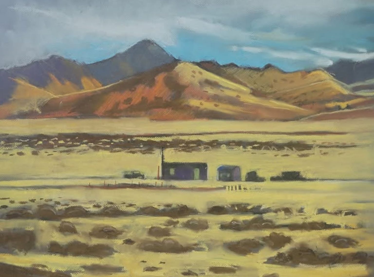 Gallery 5 - Small house in the desert. A painting of a vast, open desert landscape. The foreground is a light tan and gold field with scattered dark patches of shrubs. In the middle ground, three small, dark purple or grey structures sit alone, surrounded by a wire fence. One building has a distinctly steep roof, and another has a chimney. The structures are dwarfed by a distant range of large, rolling mountains with warm orange, yellow, and brown tones. A cloudy blue sky is above the mountains. The painting has a soft, chalky texture.