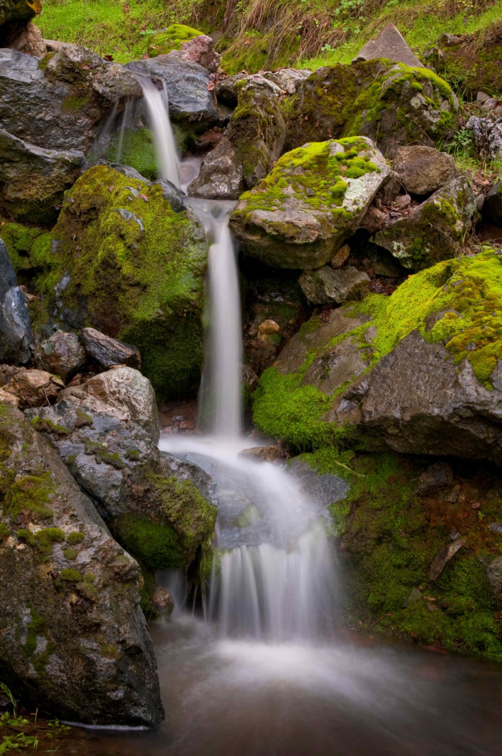 Gallery 6 - A small, multi-tiered waterfall cascades down a rocky, moss-covered slope. The water appears soft and blurry due to a long exposure, contrasting with the sharp textures of the dark, mossy rocks. Green grass and foliage are visible at the top of the frame.