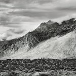 Gallery 10 - A black and white, horizontal landscape photo of a mountain range. The rugged peaks are partially obscured by low-hanging clouds. In the foreground is a low, rolling hill covered in dark, dense scrub brush.