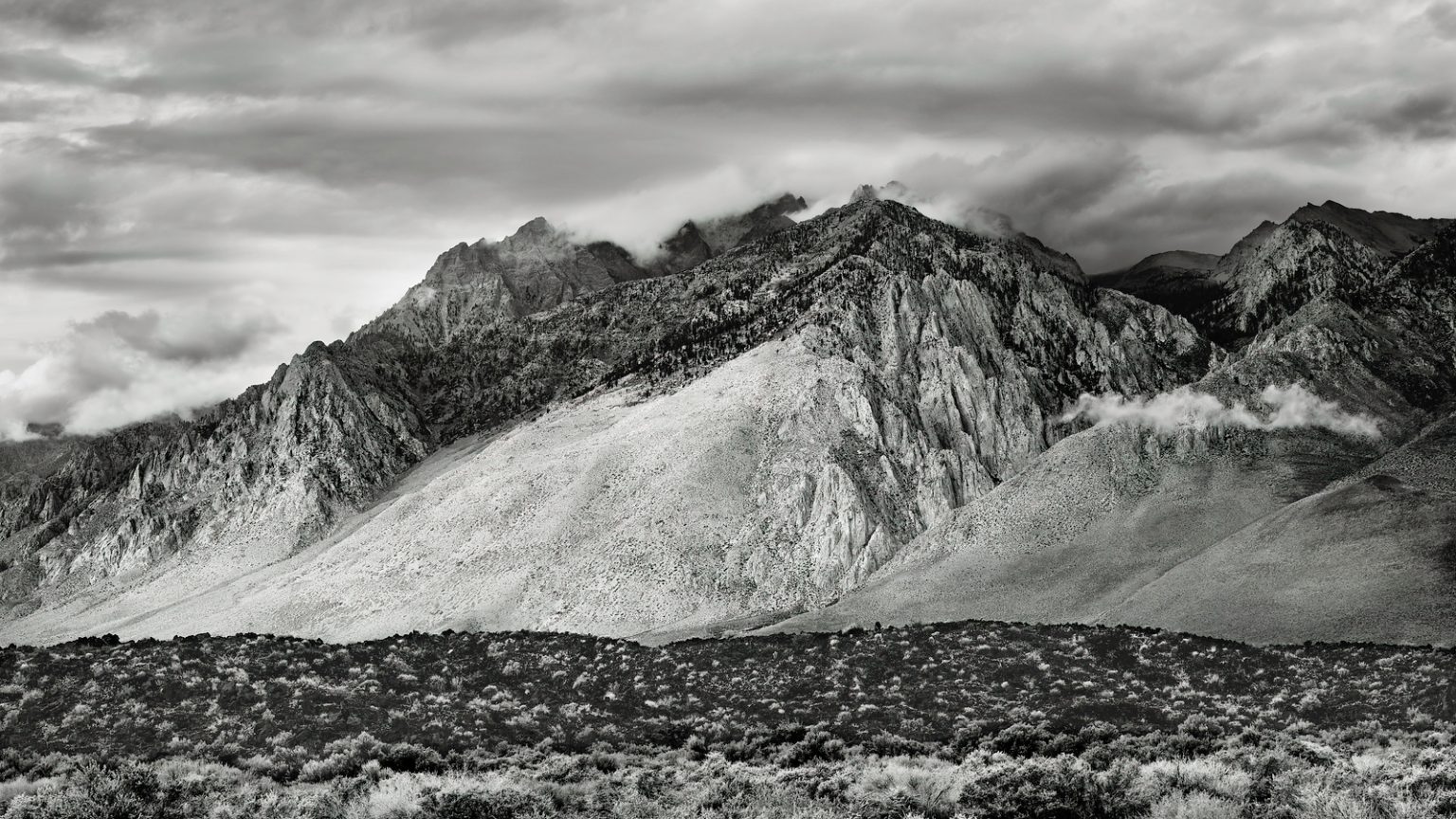 Gallery 10 - A black and white, horizontal landscape photo of a mountain range. The rugged peaks are partially obscured by low-hanging clouds. In the foreground is a low, rolling hill covered in dark, dense scrub brush.