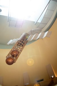 A large, suspended mixed-media sculpture by Kent Roberts, resembling a cage-like dirigible or early flying machine with fabric wings, a cylindrical body, and two copper-colored spheres within its frame, lit by bright natural light from above. A surveillance satellite made of wood, fiberglass, brass, aluminum.