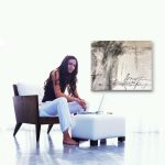 Gallery 2 - Woman in a black tank top and white pants sitting in a modern chair, using a laptop, next to a muted abstract painting with script text.