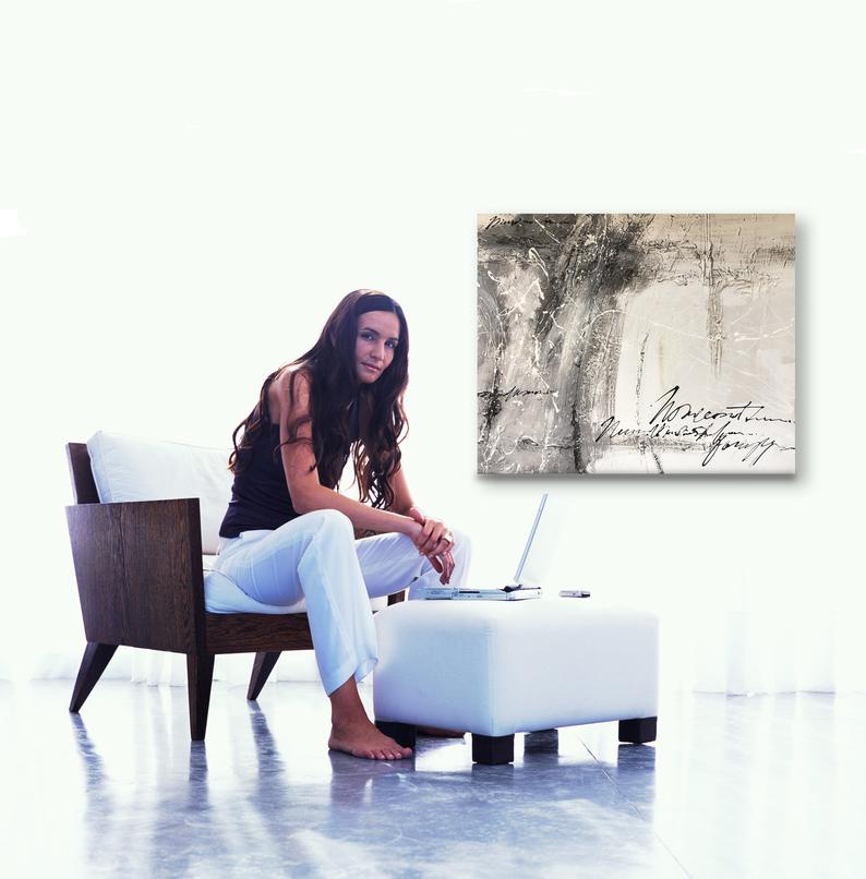 Gallery 2 - Woman in a black tank top and white pants sitting in a modern chair, using a laptop, next to a muted abstract painting with script text.