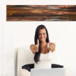 Gallery 3 - Smiling woman giving two thumbs up while sitting under a long, horizontal abstract painting with dark, earthy tones.