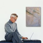 Gallery 6 - Older man in a gray turtleneck and trousers sitting on a black bench, using a laptop next to a muted abstract painting.
