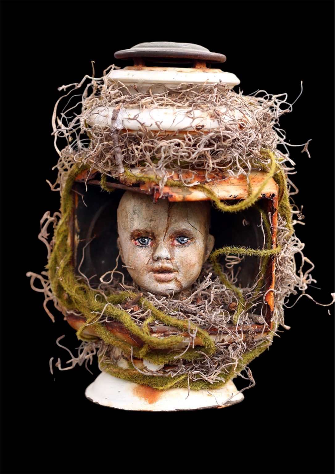 Gallery 6 - A dark and eerie mixed-media sculpture features a cracked baby doll head with blue eyes encased in an old, rusty lantern that is heavily decorated with dried moss and Spanish moss against a solid black background.