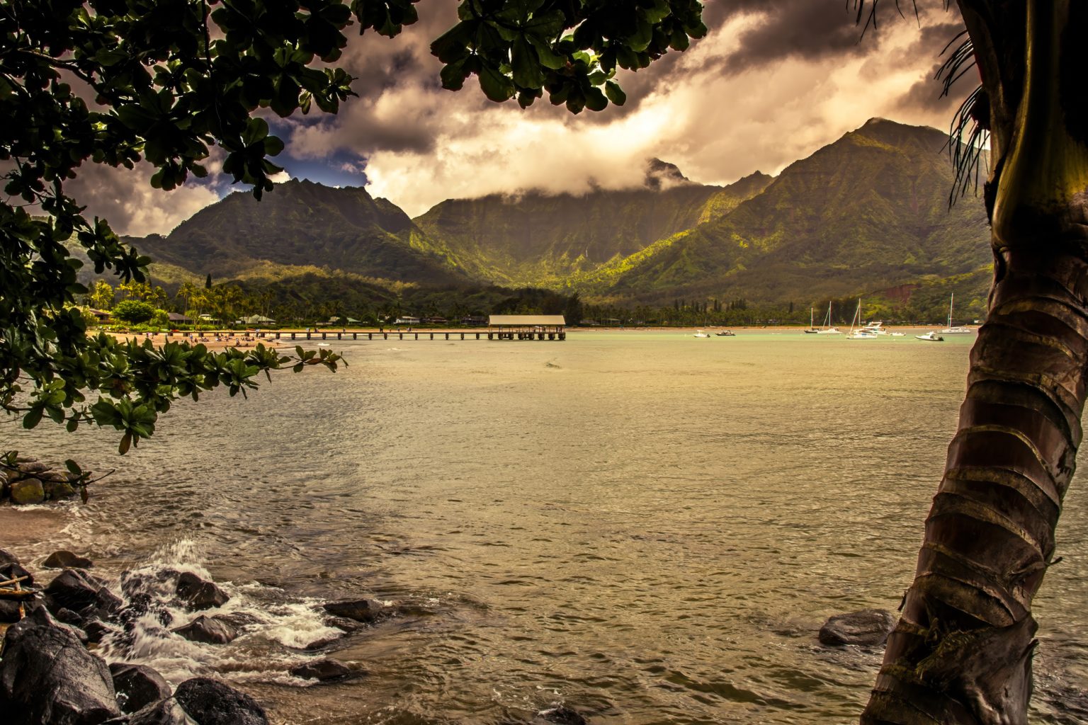 Gallery 7 - Picture of the pier at Hanalei Bay that is framed by trees on the beach and beautiful mountains.
