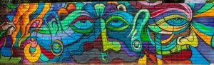 Panoramic street art mural painted on a brick wall, featuring a highly colorful, abstract, and psychedelic depiction of multiple overlapping faces and eyes in Cubist and Expressionist styles, using bright blues, greens, yellows, reds, and purples.