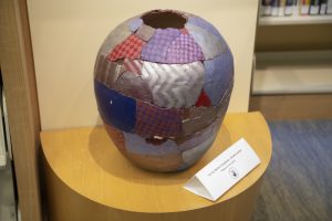 A round, ceramic pot or vessel displayed on a wooden pedestal. Its surface is covered in a colorful, irregular patchwork pattern featuring metallic silver, purple checkerboard, blue, and red segments.