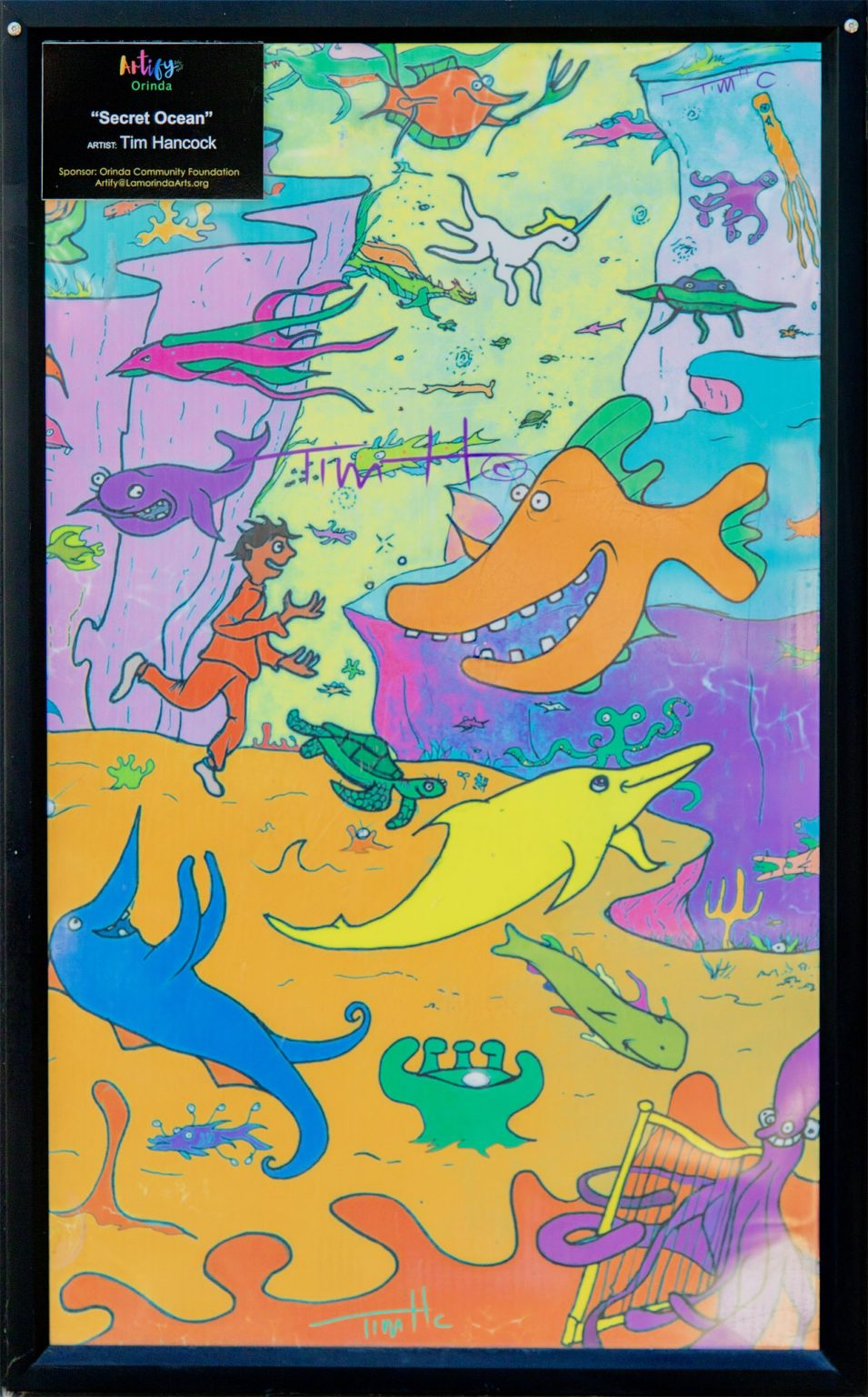 Gallery 1 - A brightly colored, cartoon-style mural on a rectangular Bigbelly trash bin showing a human figure swimming among numerous smiling and whimsical sea creatures, including a large orange fish and an octopus playing a golden harp.