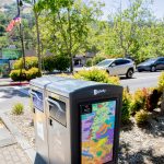 Gallery 2 - A brightly colored, cartoon-style mural on a rectangular Bigbelly trash bin showing a human figure swimming among numerous smiling and whimsical sea creatures, including a large orange fish and an octopus playing a golden harp.