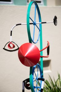 A brightly colored outdoor metal sculpture featuring two blue wheels connected by a teal belt, a large red cone, and black and white striped support legs, standing on a round concrete base.