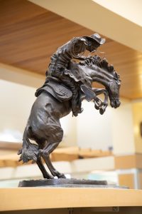 A bronze sculpture by Frederic Remington titled The Broncho Buster depicting a cowboy wearing a wide-brimmed hat and chaps, expertly riding a powerful, wildly bucking horse. The sculpture is mounted on a rectangular marble base and sits on a wooden desk or table inside a well-lit room with bookshelves visible in the background.
