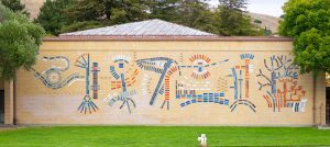 A large, colorful abstract mosaic tile mural on the beige brick wall of a building at Contra Costa College. The artwork features stylized, interconnected symbols in blue, red, orange, and white, suggesting themes of science, nature, and evolution. A grassy lawn and a road are in the foreground, with trees and hills in the background.