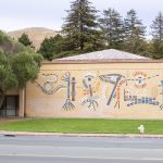Gallery 1 - A large, colorful abstract mosaic tile mural on the beige brick wall of a building at Contra Costa College. The artwork features stylized, interconnected symbols in blue, red, orange, and white, suggesting themes of science, nature, and evolution. A grassy lawn and a road are in the foreground, with trees and hills in the background.
