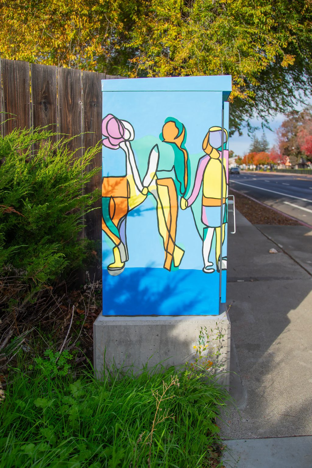 Gallery 1 - A utility box painted with a bright, abstract mural of a family, featuring yellow, orange, green, and purple figures of a tall parent, a shorter parent, and a child holding hands. The sun flares over the top right corner of the box, which sits on a concrete base next to a sidewalk.