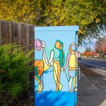 Gallery 1 - A utility box painted with a bright, abstract mural of a family, featuring yellow, orange, green, and purple figures of a tall parent, a shorter parent, and a child holding hands. The sun flares over the top right corner of the box, which sits on a concrete base next to a sidewalk.