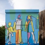 Gallery 2 - A utility box painted with a bright, abstract mural of a family, featuring yellow, orange, green, and purple figures of a tall parent, a shorter parent, and a child holding hands. The sun flares over the top right corner of the box, which sits on a concrete base next to a sidewalk.