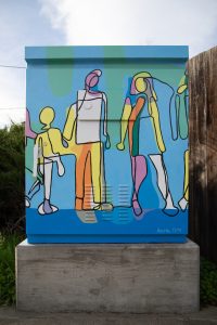 A utility box painted with a bright, abstract mural of a family, featuring yellow, orange, green, and purple figures of a tall parent, a shorter parent, and a child holding hands. The sun flares over the top right corner of the box, which sits on a concrete base next to a sidewalk.