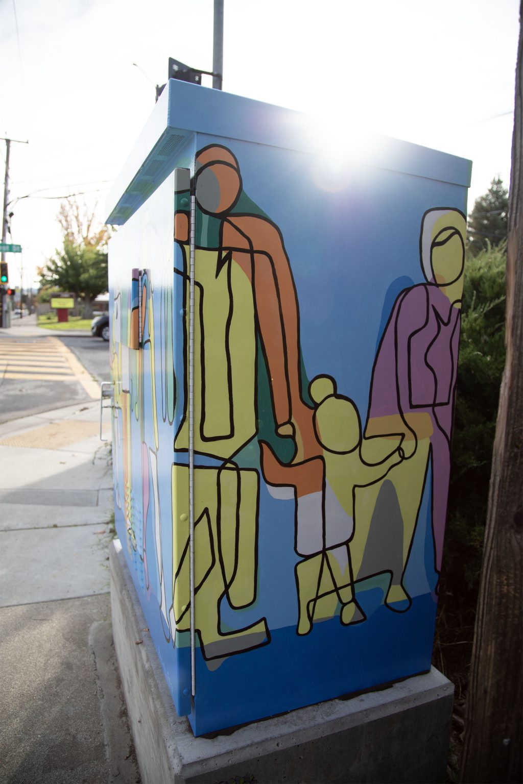 Gallery 4 - A utility box painted with a bright, abstract mural of a family, with yellow, orange, green, and purple figures of a tall parent, a shorter parent, and a child holding hands. The sun flares over the top right corner of the box.