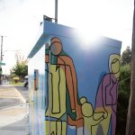 Gallery 4 - A utility box painted with a bright, abstract mural of a family, with yellow, orange, green, and purple figures of a tall parent, a shorter parent, and a child holding hands. The sun flares over the top right corner of the box.