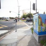 Gallery 3 - A utility box painted with a bright, abstract mural of a family, with yellow, orange, green, and purple figures of a tall parent, a shorter parent, and a child holding hands. The sun flares over the top right corner of the box.