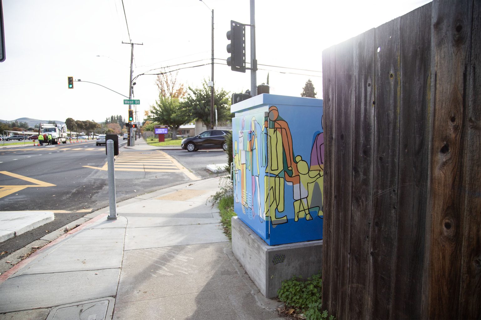 Gallery 3 - A utility box painted with a bright, abstract mural of a family, with yellow, orange, green, and purple figures of a tall parent, a shorter parent, and a child holding hands. The sun flares over the top right corner of the box.