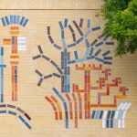 Gallery 2 - Close up of a large, colorful abstract mosaic tile mural on the beige brick wall of a building at Contra Costa College. The artwork features stylized, interconnected symbols in blue, red, orange, and white, suggesting themes of science, nature, and evolution. A grassy lawn and a road are in the foreground, with trees and hills in the background.